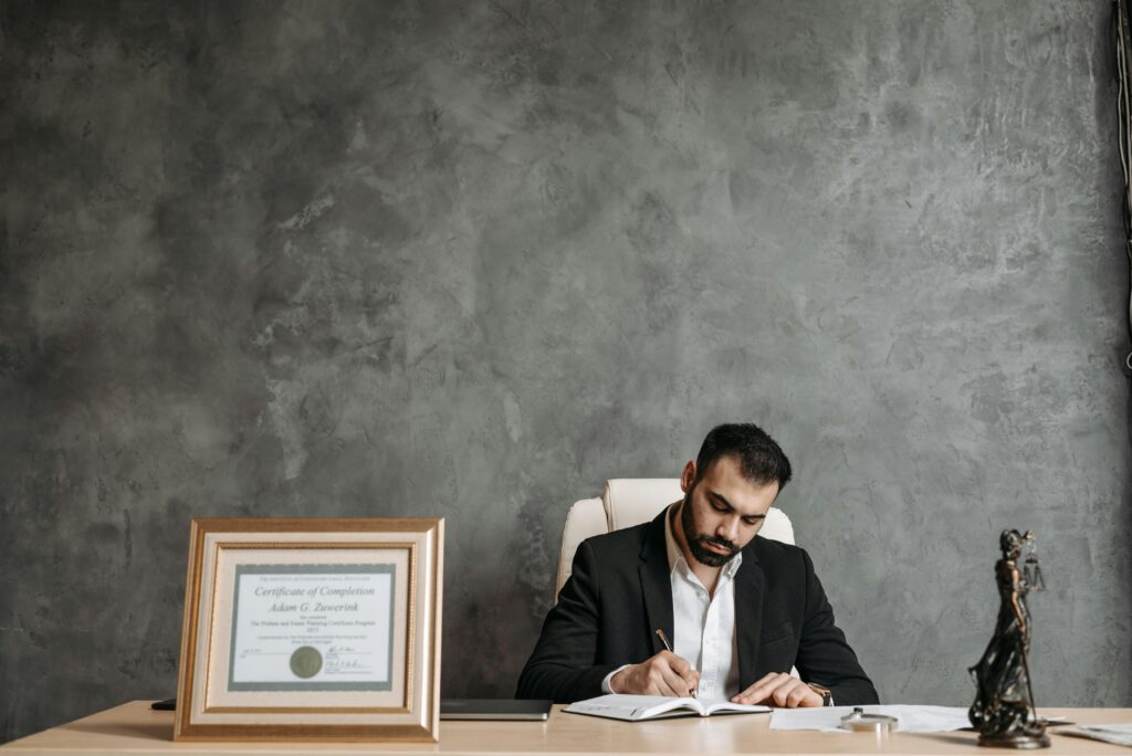 A business professional writing at a desk with a framed certificate and justice statue.