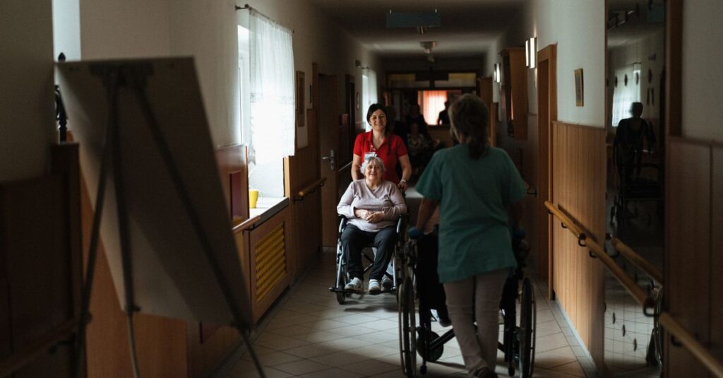 Caregiver assisting elderly woman in a wheelchair through a retirement home corridor.