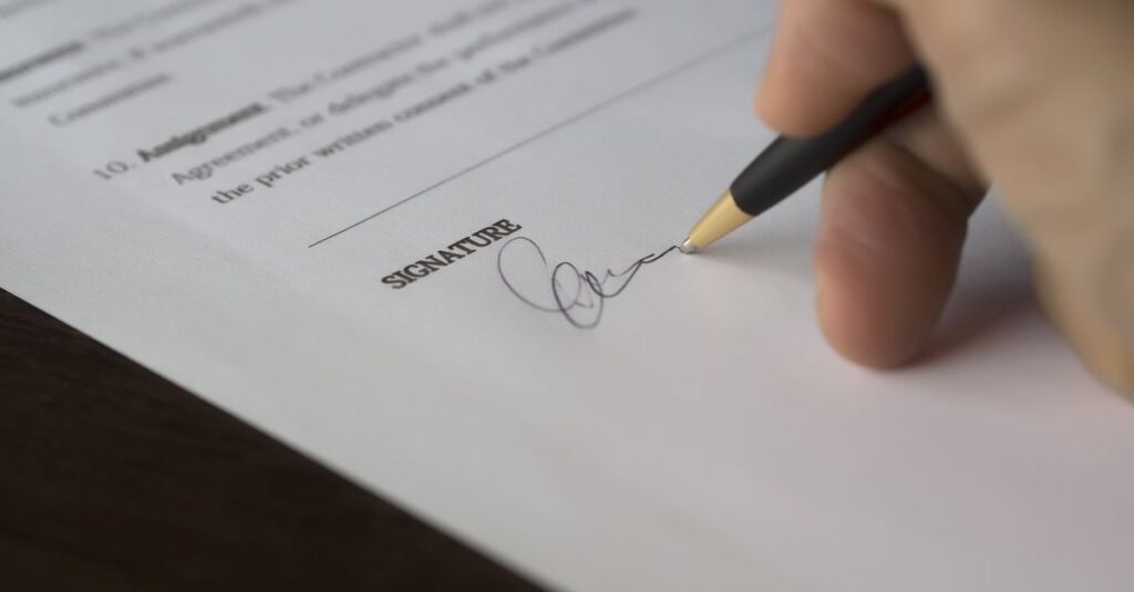 A hand signs a formal contract with a pen on a wooden desk.