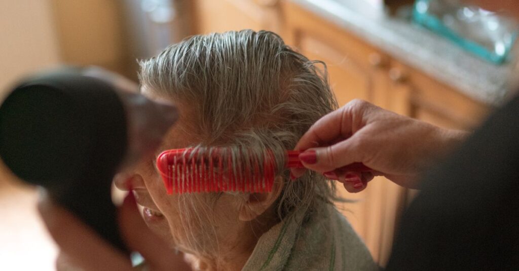 A caregiver assists a senior adult with hair drying and combing at home.