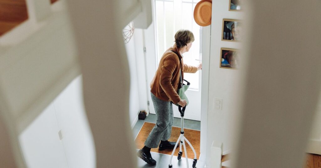 Senior woman using rollator indoors near doorway, preparing to leave home.