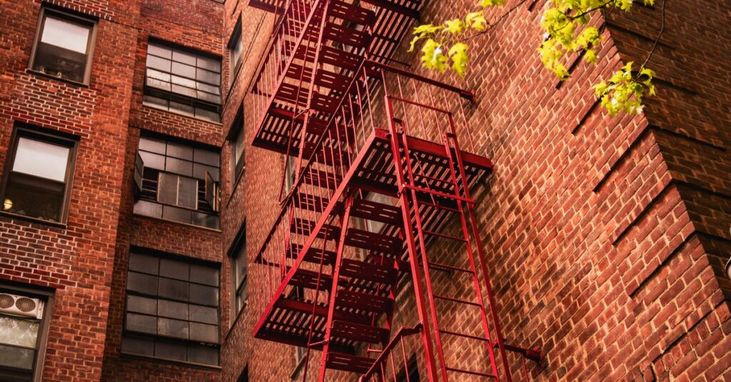 Red fire escape on a classic New York brick building under spring foliage.