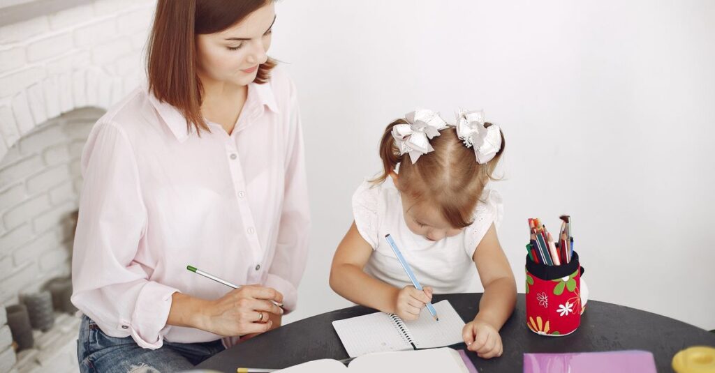 From above small girl drawing with pencil in notepad while mother sitting near at round table