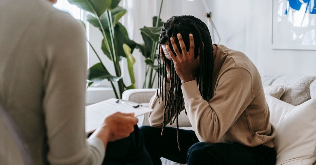 Anxious African American male with hands on head sitting on sofa near crop female psychologist with clipboard during appointment in office