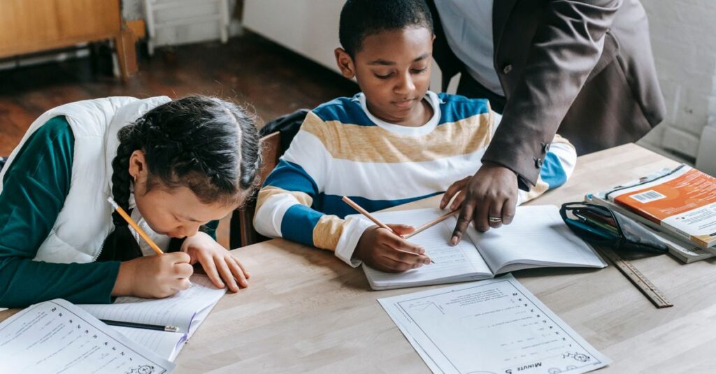 High angle of crop unrecognizable black female teacher explaining task to focused little schoolboy sitting at desk near attentive Asian classmate writing in notebook