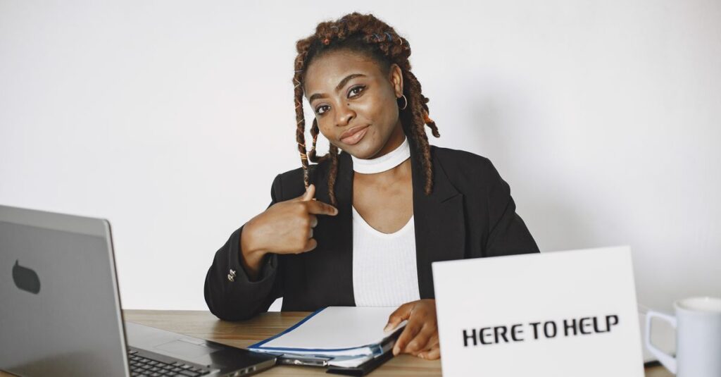 A friendly businesswoman in an office setting, ready to assist with a sign reading 'Here to Help'.