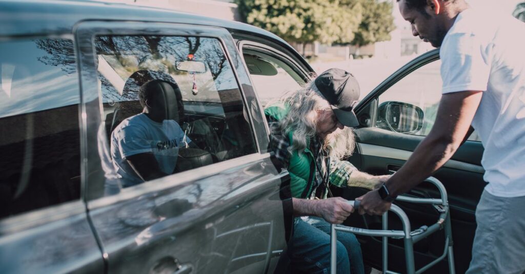 Two men help an elderly person with a walker get out of a car outdoors.