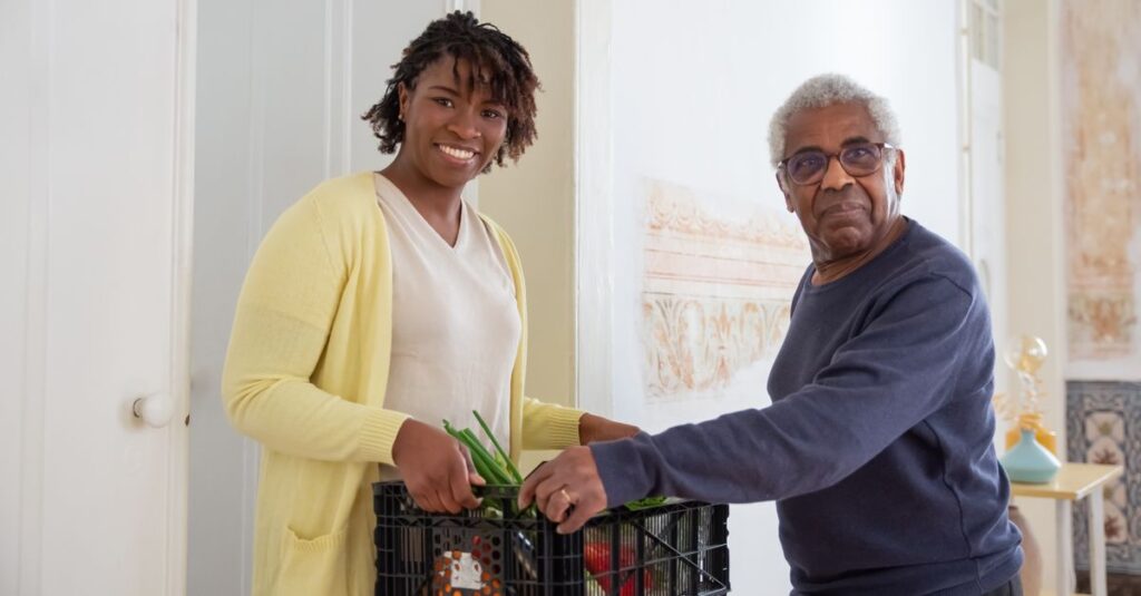 Smiling woman assists a senior man with food delivery indoors, promoting community support and care.