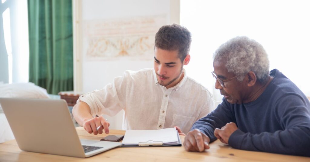 Young man teaches elderly gentleman to use technology at home.
