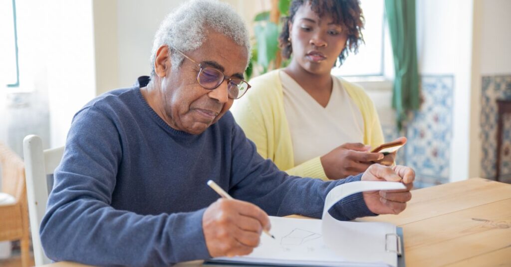 A senior man writing on papers with a caregiver nearby indoors in a supportive setting.