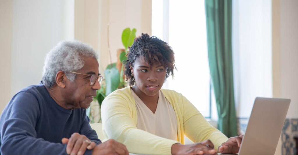 Young woman assists an elderly man with laptop indoors, highlighting intergenerational tech support.