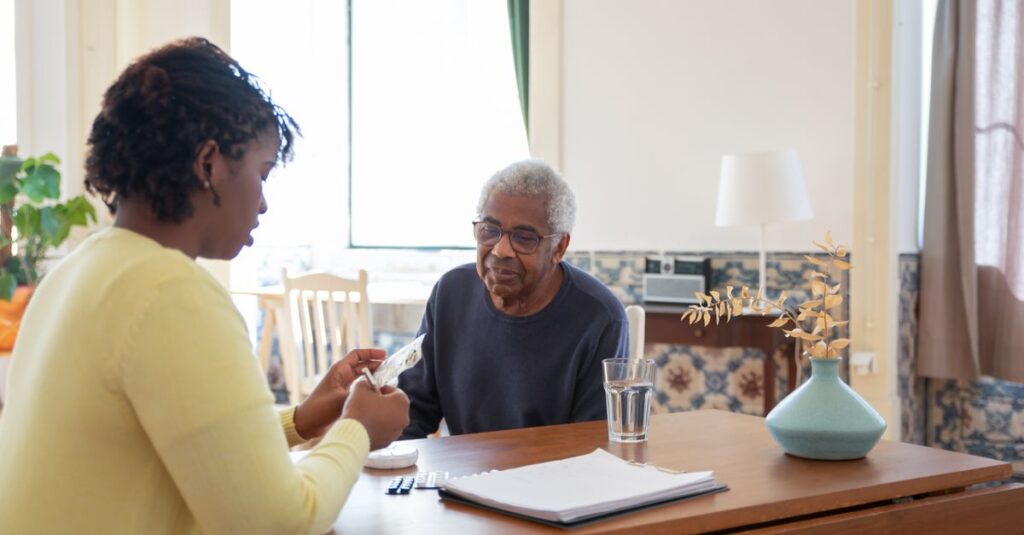 A caregiver assists an elderly man with medication in a warm, sunlit living room.