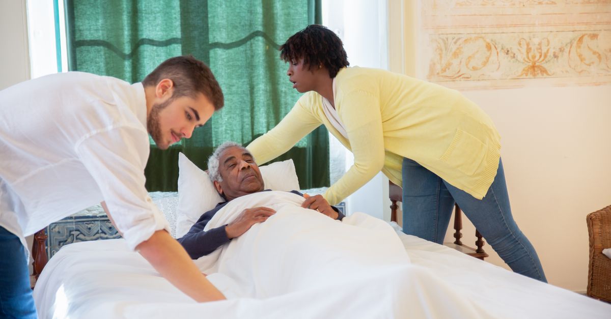 Caregivers supporting an elderly man lying in bed, providing assistance and comfort.