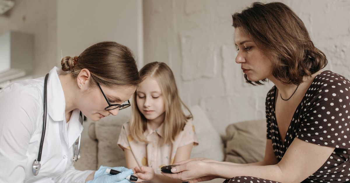 A pediatrician checks a child's blood sugar levels while her mother assists at home.