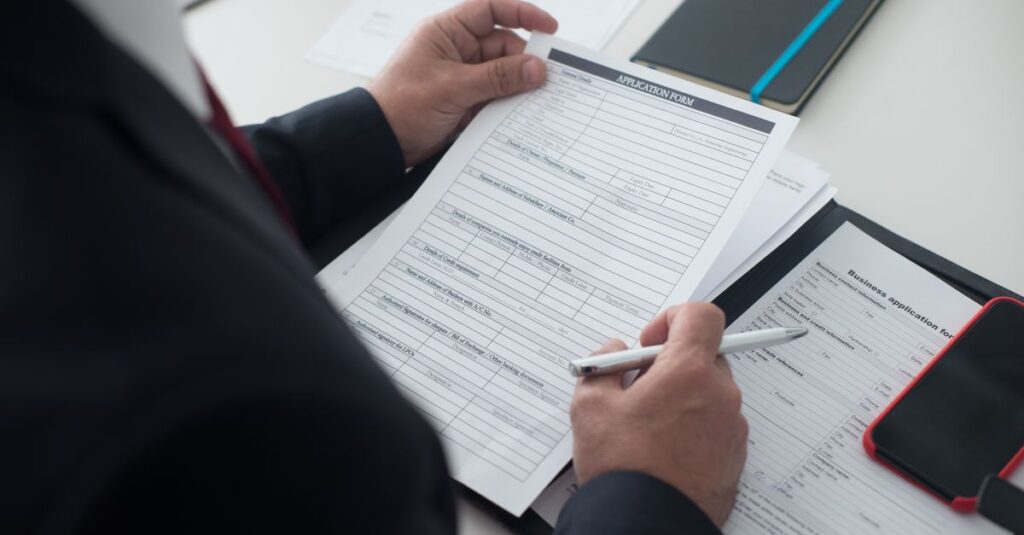 A businessman in a black suit reviews a printed application form with a pen on his desk.