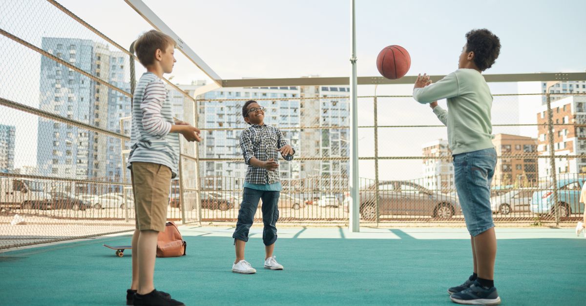 Three boys playing basketball on an outdoor court surrounded by city buildings.