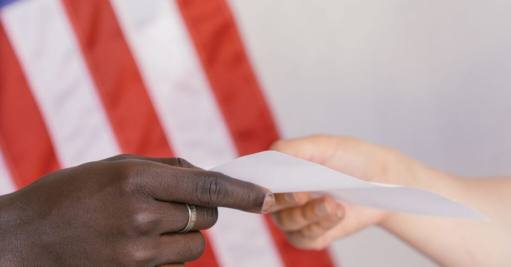 Two hands exchanging a paper in front of a blurred American flag background.