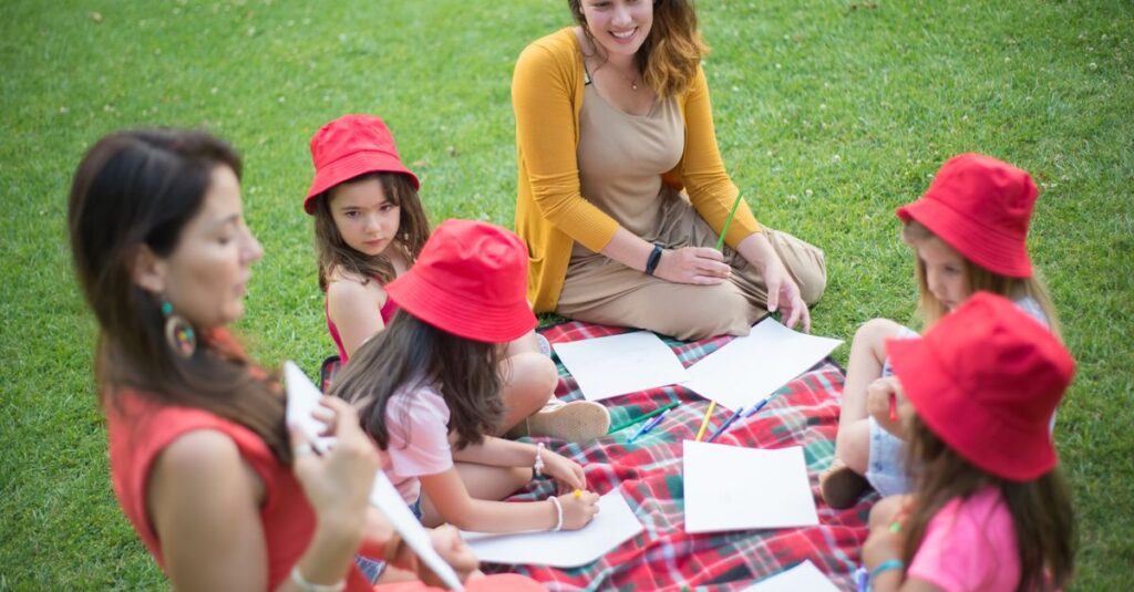 Children and teacher sitting on a blanket in a park, engaged in art activities.