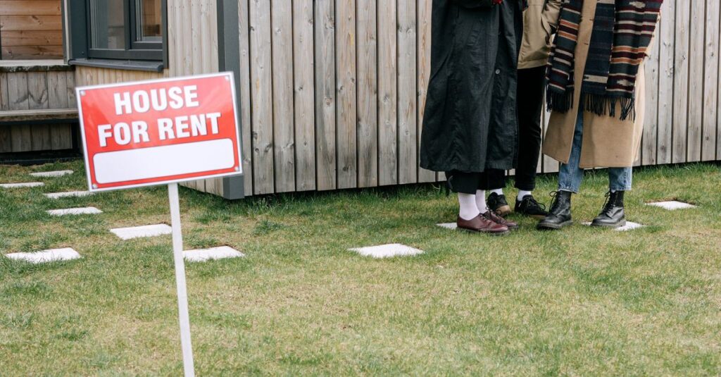 A group of three people reviewing a tablet near a 'House for Rent' sign outside.